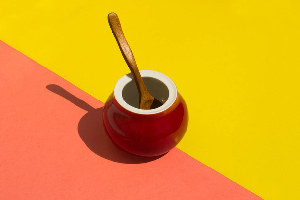 Red ceramic pot with a wooden spoon on a yellow-red background, it has a diagonal and shadows. - Valokuva, kuva