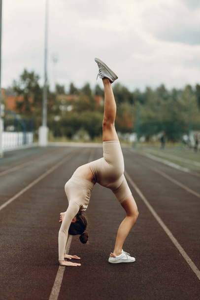 A beautiful female athlete makes a bridge on her hands in sports clothes. Stretching at the stadium on a treadmill. - 写真・画像