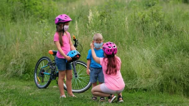Dos niñas con cascos rosas y máscaras médicas. Ayudan a su hermano menor a ponerse un casco de bicicleta. Prevención de la infección por coronavirus
. - Imágenes, Vídeo