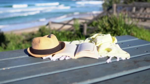 Libro, scialle e cappello sul tavolo con onde blu dell'oceano sullo sfondo. - Filmati, video