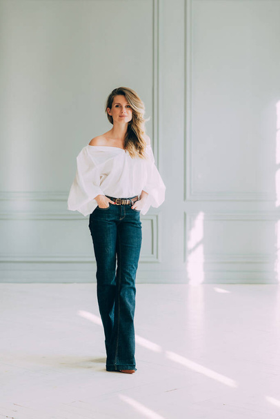 Mooi elegant sexy meisje in een witte blouse met prachtige mouwen en flared jeans in een kamer gevuld met de zon. Schoonheid en mode stijl. Zachte selectieve focus. - Foto, afbeelding