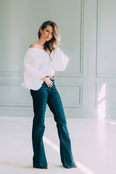 Beautiful elegant sexy girl in a white blouse with magnificent sleeves and flared jeans in a room filled with the sun. Beauty and fashion style. Soft selective focus. - Foto, Imagen