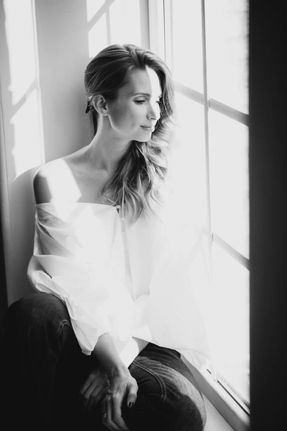 Beautiful elegant sexy girl with chubby lips girl in a white blouse with voluminous sleeves and jeans on the window in the sun. Black and white art photo. Soft selective focus. - Foto, Bild