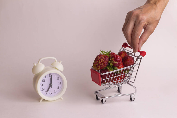 Die weibliche Hand hält einen Mini-Korb mit Erdbeeren und einer Retro-Uhr auf weißem Hintergrund. Erntezeitkonzept im Garten. Einkaufen im Online-Shop. Hauszustellung. - Foto, Bild