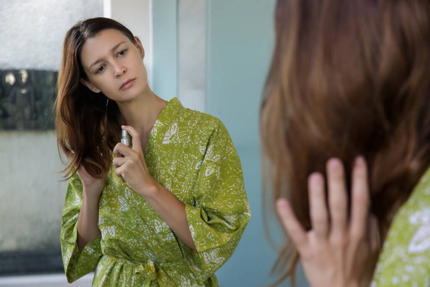 Péče o vlasy. Žena nanášející olej na vlasy končí v koupelně před zrcadlem. Rozdělit vlasy tipy, suché vlasy nebo koncepce ochrany proti slunci. - Fotografie, Obrázek