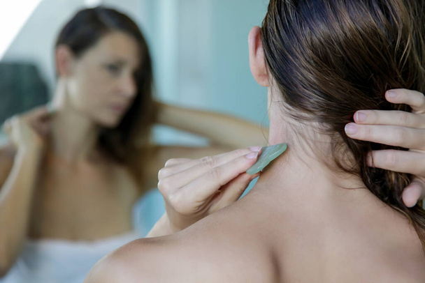 Mujer joven está haciendo Gua Sha masaje facial en el baño delante del espejo. Gua Sha piedra de jade en el tratamiento en casa
. - Foto, Imagen