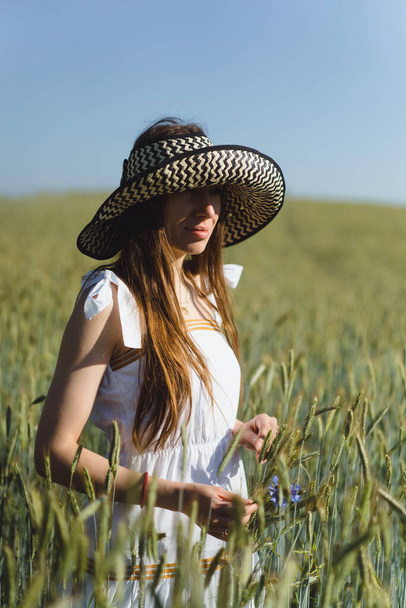 young beautiful woman with long hair in a white dress and hat among the yellow field of rye and wheat. - Foto, Bild