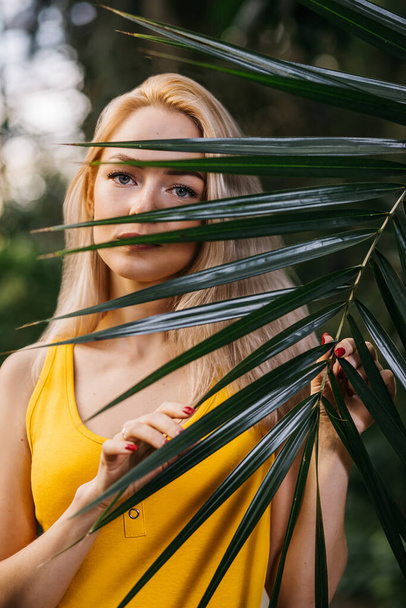 Beautiful girl with natural makeup, blonde hair standing in rainforest among exotic trees. Closeup portrait of magnificent woman in yellow swimsuit among tropical plants. Focus on beautiful palm leaves - Photo, Image