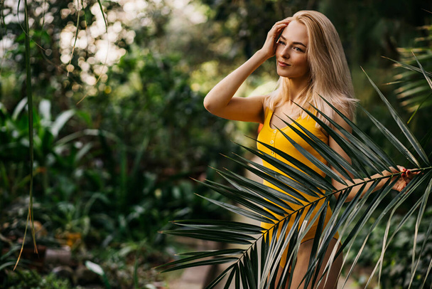 Charming caucasian woman with nude makeup and blonde hair, posing in rainforest, gentle touching face, wearing yellow swimsuit. Vacation, summer, skin care concept. - Foto, Imagem