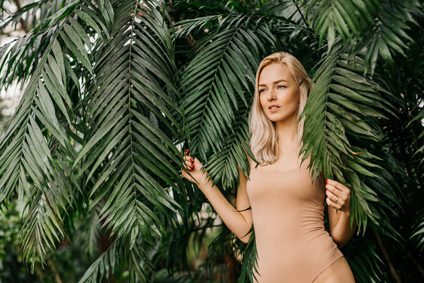 Portrait of happy lady with natural nude makeup, perfect skin, blonde hair. Beautiful woman hiding behind palm leaves and look down. Cosmetic, wellness, purity, skincare, spa concept. - Фото, изображение