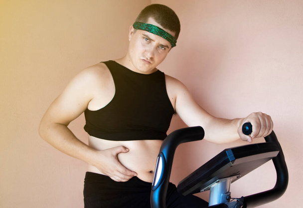 Homem gordo alegre em um traje engraçado está envolvido em uma bicicleta estacionária. O conceito de problemas com o peso, perder peso, praticar esportes e motivação para alcançar um corpo magro
. - Foto, Imagem