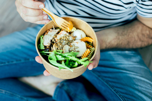 Eating healthy lunch bowl. Chicken, pasta fusilli, capers, mix greens, vegetables and sunflower seeds in paper bowl in man' s hands. Food delivery, dieting, detox, vegetarian food concept - Photo, Image