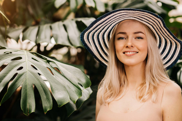 Lovely amazed blonde lady wearing sun hat, flesh color swimsuit, looking into distance, standing with raised hands on tropical plants background. Summer vacation, facial expressions, emotion concept. - Foto, Imagem