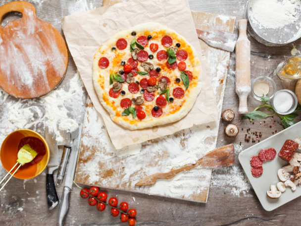 Top overhead view cooking pizza with ingredients, tomatoes, salami, flour, dough basil, spices, mozzarella and mushrooms on wooden tabletop - Photo, image