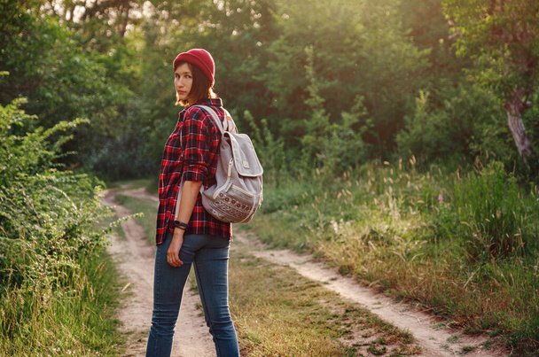 A fiatal hipszter lány élvezi a naplementét az erdőben. Utazó nő hátizsákkal. Élvezem a naplementét. Pihenés a parkban napkeltekor. Trekking-koncepció - Fotó, kép