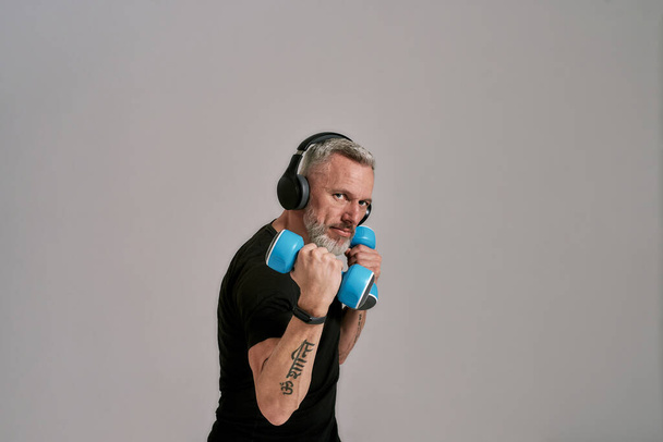 Middle aged muscular man in black t shirt and headphones looking at camera holding blue dumbbells, posing in studio over grey background - Zdjęcie, obraz