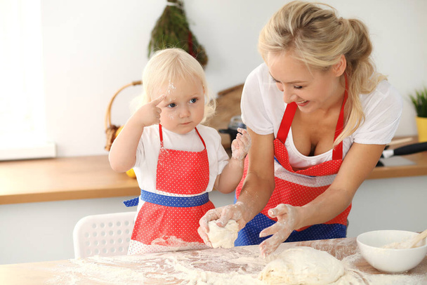 Little girl and her blonde mom in red aprons playing and laughing while kneading the dough in kitchen. Homemade pastry for bread, pizza or bake cookies. Family fun and cooking concept - Photo, Image