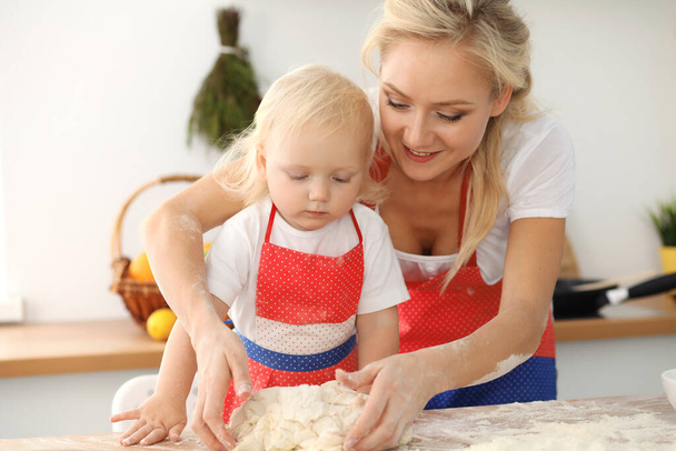 Mała dziewczynka i jej blond mama w czerwonych fartuchach bawią się i śmieją podczas ugniatania ciasta w kuchni. Domowe ciasto na chleb, pizzę lub ciasteczka. Rodzinna zabawa i koncepcja gotowania - Zdjęcie, obraz