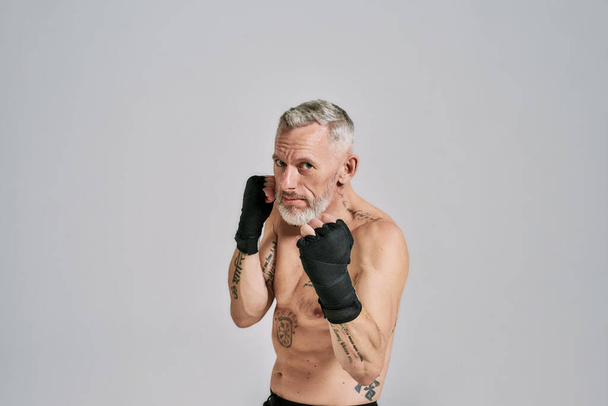 Half naked middle aged athletic man, kickboxer looking at camera while boxing, practicing punches in studio over grey background - Photo, Image