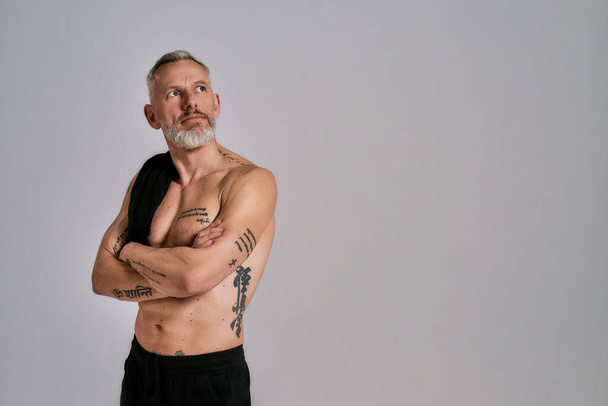 Middle aged muscular man took off black t shirt, showing his body, looking aside while posing in studio over grey background - Foto, afbeelding