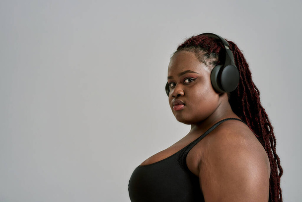 Plump, plus velikost africké americké ženy ve sportovním oblečení a sluchátka při pohledu do kamery, stojící ve studiu nad šedým pozadím. Pojetí sportu, zdravý životní styl, tělo pozitivní, rovnost - Fotografie, Obrázek