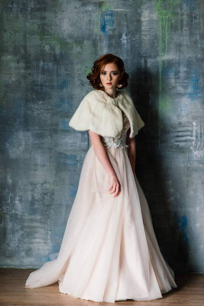 Portrait of red haired girl wearing wedding dress against a white and grey studio background. - Foto, imagen