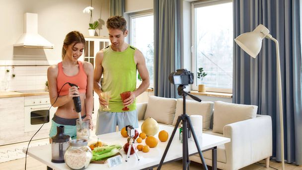Capture it all. Young couple recording video blog or vlog about healthy nutrition on camera at home. Man and woman showing ingredients and explaining how to prepare, standing in the kitchen - Fotó, kép