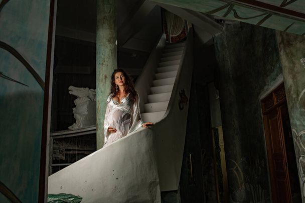 low angle view of beautiful smiling young pregnant woman in white peignoir posing on staircase  - Foto, immagini