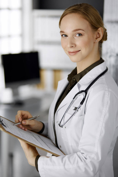 Cheerful smiling female doctor using clipboard in clinic. Portrait of friendly physician woman at work. Perfect medical service in hospital. Medicine concept - Фото, зображення