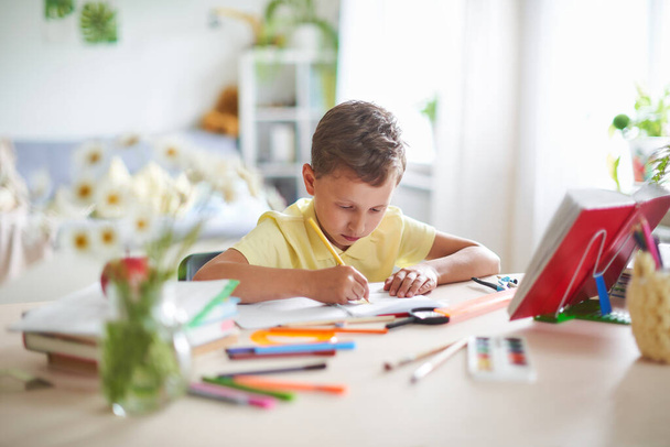 capable boy 7 years old, enthusiastically writes and draws at table. boy does his homework. Home schooling. Do your math homework at HOME, at your Desk. On table are school supplies. - Photo, image