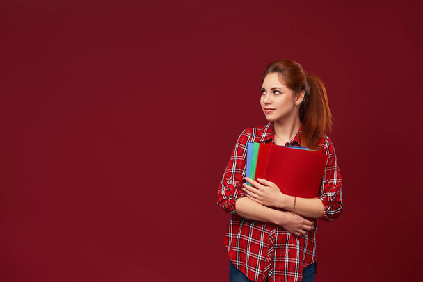 teenage girl is isolated on a red background, holding colorful folders in her hands. attractive Caucasian student dressed in checked shirt is ready for training. Concept education. time of exams. - Photo, Image