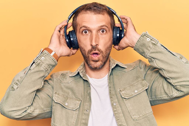 Młody przystojny mężczyzna słuchający muzyki przy użyciu słuchawek przerażony i zdumiony otwartymi ustami dla zaskoczenia, niedowierzanie twarz  - Zdjęcie, obraz