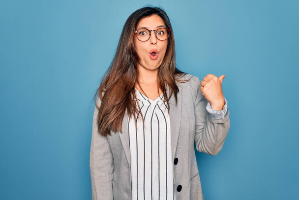 Nuori latino liiketoiminnan nainen yllään lasit seisoo sininen eristetty tausta Yllättynyt osoittaa sormella sivulle, avoin suu hämmästynyt ilme. - Valokuva, kuva