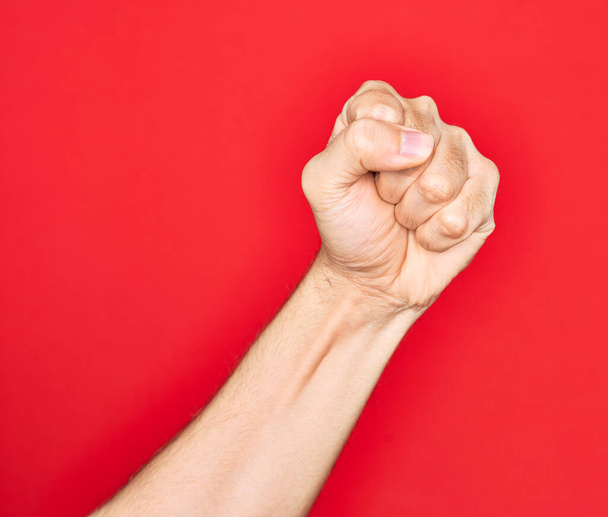 Mano de joven caucásico mostrando dedos sobre fondo rojo aislado haciendo protesta y gesto de revolución, puño expresando fuerza y poder
 - Foto, Imagen