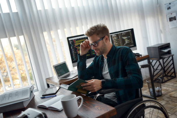 Successful people never stop learning. Side view of young male web developer in a wheelchair reading a book and adjusting his eyeglasses while sitting in the modern office - Foto, immagini