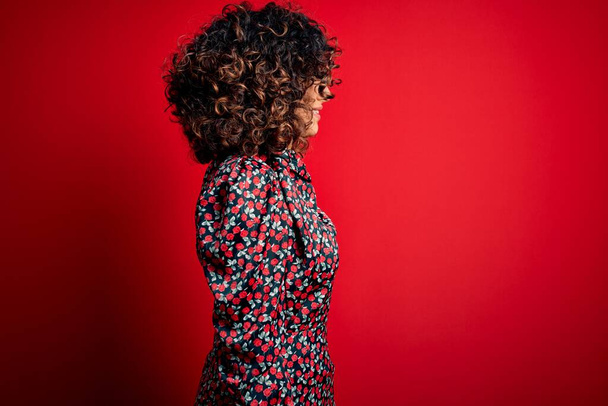 Young beautiful curly arab woman wearing casual floral dress standing over red background looking to side, relax profile pose with natural face with confident smile. - Fotoğraf, Görsel