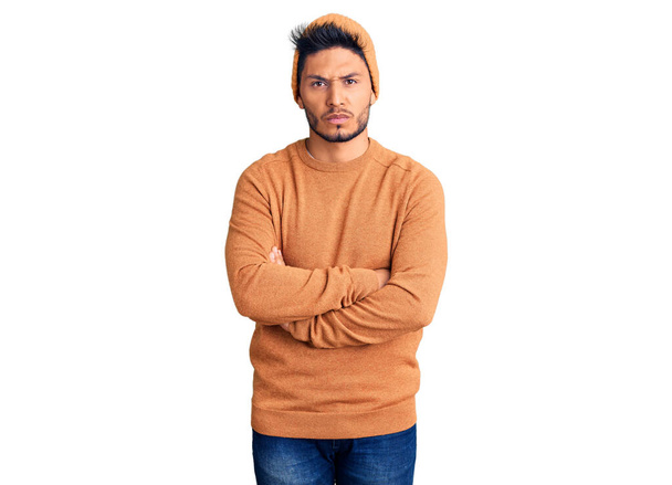 冬のセーターとウールの帽子を身に着けているハンサムなラテン系アメリカ人の若者は懐疑的で緊張し、両腕を組んで顔に不承認の表現。否定的な人.  - 写真・画像