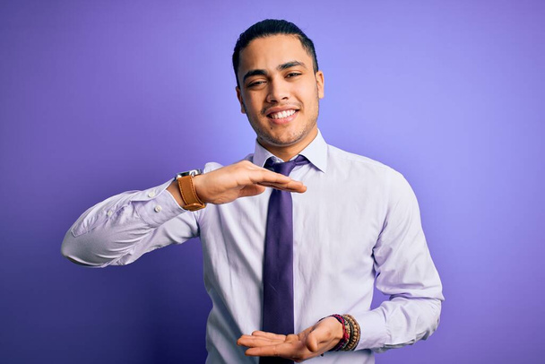 Young brazilian businessman wearing elegant tie standing over isolated purple background gesturing with hands showing big and large size sign, measure symbol. Smiling looking at the camera. Measuring concept. - Fotografie, Obrázek