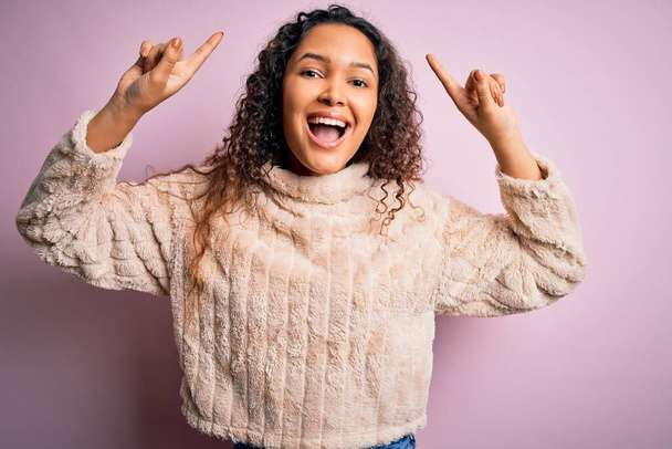 Jonge mooie vrouw met krullend haar dragen casual trui staan over roze achtergrond lachend verbaasd en verrast en wijzen omhoog met vingers en opgeheven armen. - Foto, afbeelding