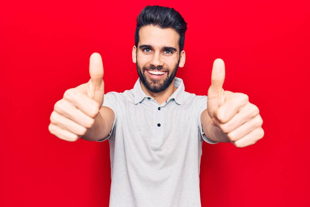 Young handsome man with beard wearing casual polo approving doing positive gesture with hand, thumbs up smiling and happy for success. winner gesture.  - Photo, Image