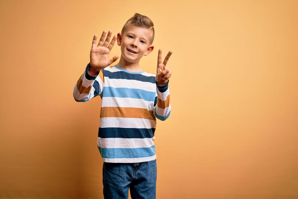 Jovem garoto caucasiano com olhos azuis vestindo camisa listrada colorida sobre fundo amarelo mostrando e apontando para cima com os dedos número sete enquanto sorri confiante e feliz
. - Foto, Imagem