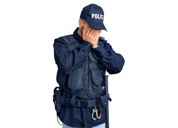 Jeune homme hispanique portant un uniforme de police avec une expression triste couvrant le visage avec les mains tout en pleurant. concept de dépression.  - Photo, image