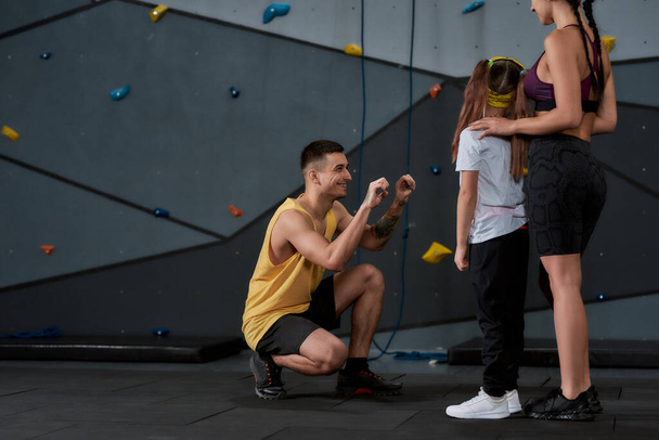 Niña activa que va a escalar, instructor masculino y femenino que ayuda al niño, de pie contra la pared de escalada de entrenamiento artificial
 - Foto, imagen