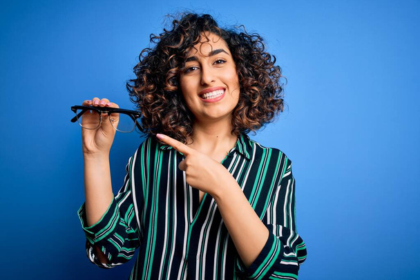 Молодая красивая оптическая арабская женщина держит очки зрения на изолированном голубом фоне очень счастливо указывая рукой и пальцем
 - Фото, изображение