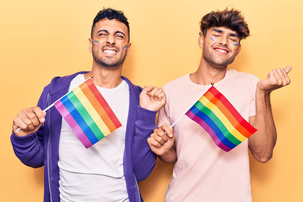 jovem gay casal segurando arco-íris lgbtq bandeiras gritando orgulhoso, celebrando vitória e sucesso muito animado com levantou braço
  - Foto, Imagem