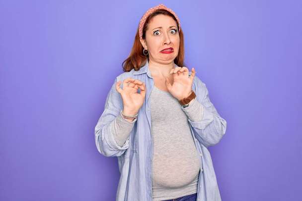 Νεαρή όμορφη κοκκινομάλλα έγκυος γυναίκα περιμένει μωρό πάνω από απομονωμένο μωβ φόντο αηδιασμένη έκφραση, δυσαρεστημένος και φοβισμένος κάνει αηδία πρόσωπο επειδή αντίδραση αποστροφή. Με τα χέρια ψηλά - Φωτογραφία, εικόνα