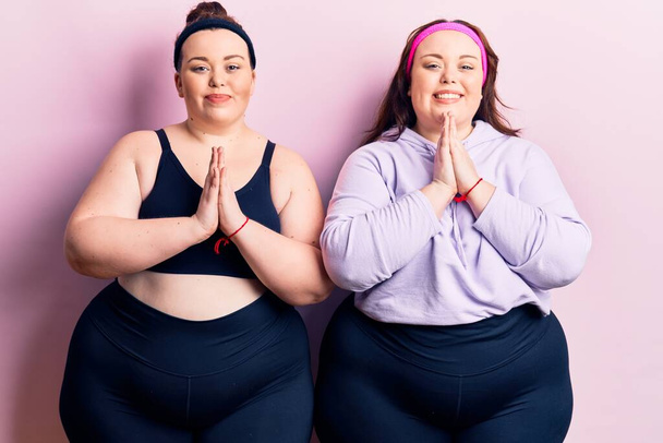 Young plus size twins wearing sportswear praying with hands together asking for forgiveness smiling confident.  - Photo, Image