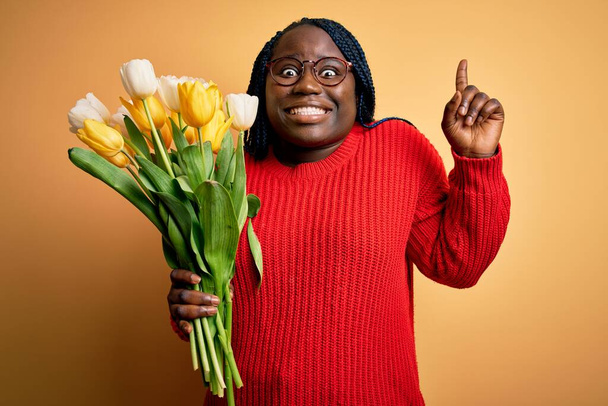 Joven afroamericana mujer de talla grande con trenzas sosteniendo ramo de tulipanes amarillos flor sonriendo asombrado y sorprendido y apuntando hacia arriba con los dedos y los brazos levantados
. - Foto, imagen
