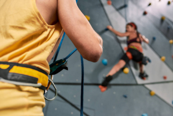 Обрезанный вид мужчины-инструктора, держащего верёвку, в то время как женщина в технике безопасности и тренировке ремней безопасности на искусственной скалолазающей стене
 - Фото, изображение