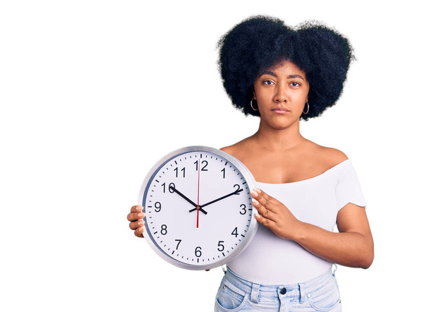 Νεαρή Αφρο-Αμερικανίδα που κρατάει μεγάλο ρολόι σκεπτόμενη στάση και νηφάλια έκφραση δείχνοντας αυτοπεποίθηση  - Φωτογραφία, εικόνα